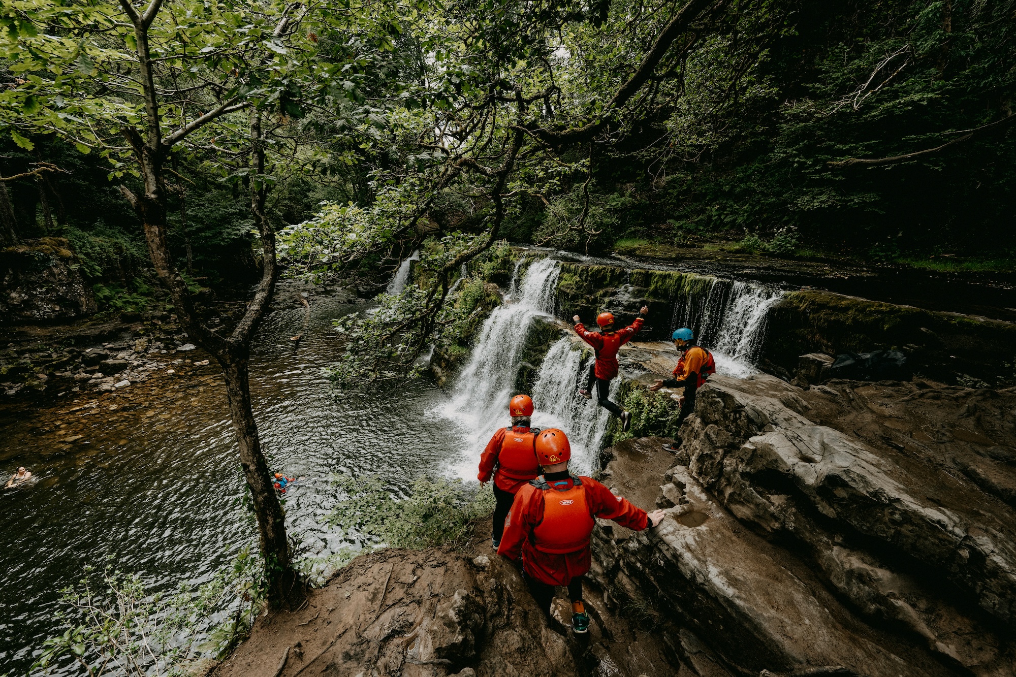Group in orange dry suits exploring a gorge waterfall in lush green woodland