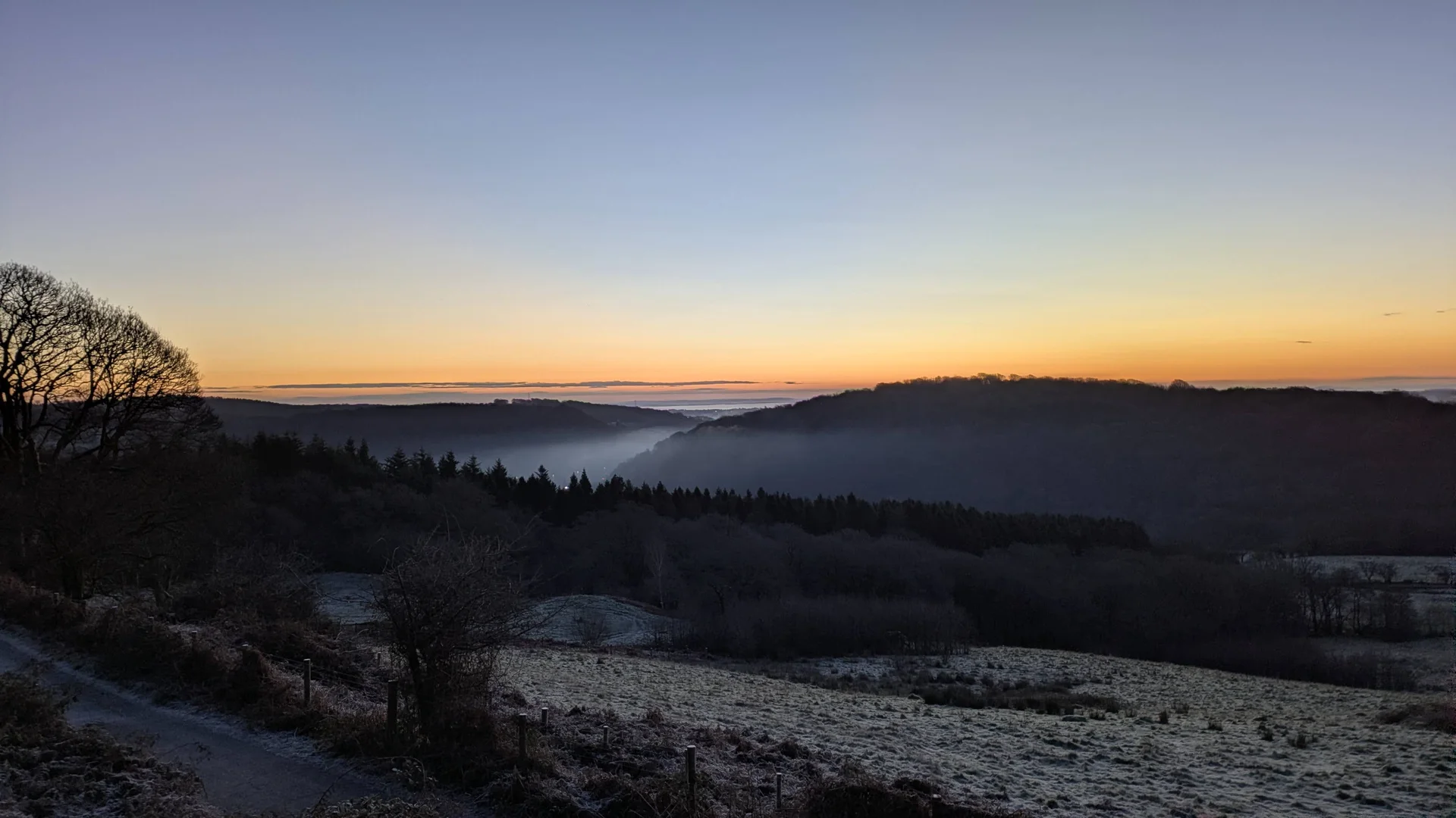 Dawn light glowing orange on the horizon over a misty valley, frosty fields in the foreground