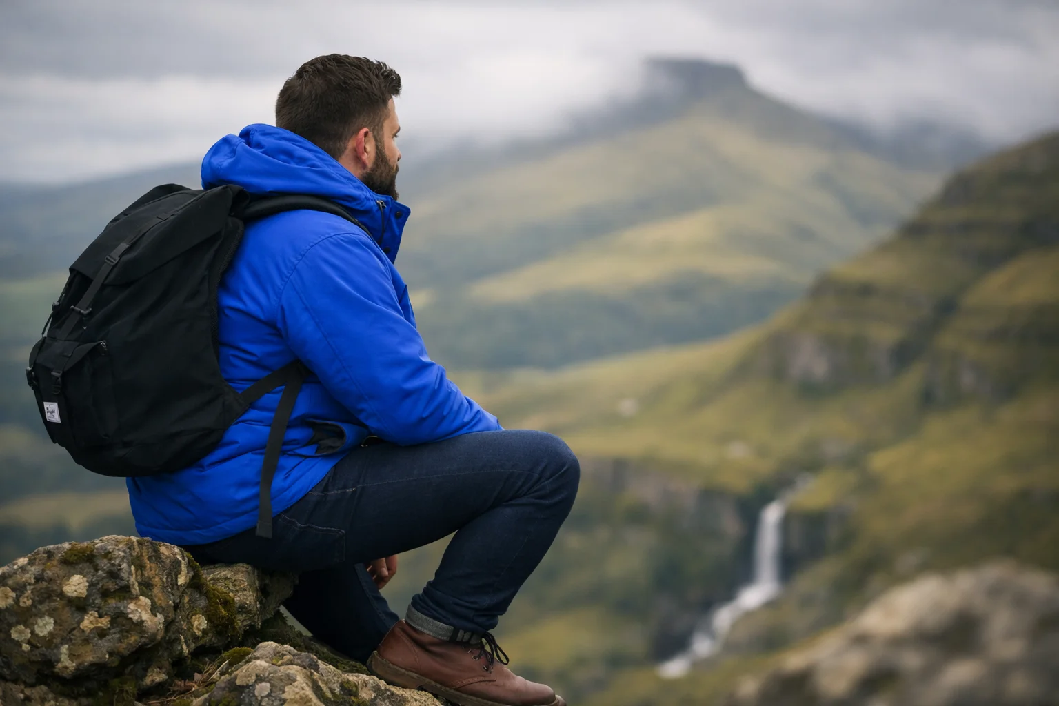 A walker in a blue jacket sitting on a rocky outcrop above a wide valley